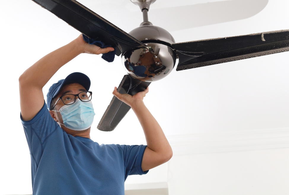 A professional technician from J.A. Bertsch Heating & Cooling in Coeur d'Alene, ID, wearing a mask and blue uniform, cleans and services a ceiling fan. The technician ensures proper maintenance to enhance the fan's performance and energy efficiency. Ceiling fan maintenance, fan cleaning service, electrical repairs Coeur d'Alene, home maintenance Post Falls, HVAC services Hayden, residential electrical service Rathdrum, professional electrician Sandpoint, fan troubleshooting Athol, energy efficiency, home electrical safety.