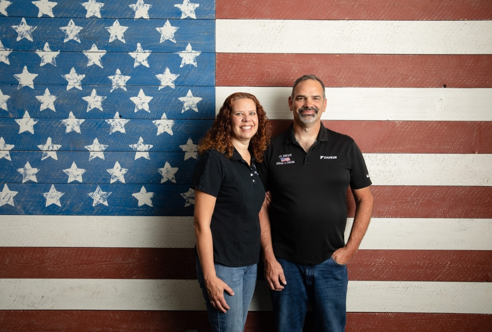 Two smiling J.A. Bertsch Heating, Cooling & Electrical employees standing in front of an American flag, representing trusted electrical services in Coeur d’Alene including electrical repairs, wiring upgrades, outlet installation, electrical panel replacement, and lighting installation. Call us today for help with electrical services, residential electrical services, commercial electrical services, licensed electricians, certified electricians near me, electrical repairs, home electrical repair, electrical troubleshooting, electrical upgrades, circuit breaker replacement, electrical panel replacement, wiring repair, electrical rewiring, whole home rewiring, electrical inspections, lighting installation, indoor lighting repair, outdoor lighting installation, ceiling fan wiring, outlet installation, GFCI outlet repair, surge protection installation, EV charger installation, generator installation, emergency electrical services, 24/7 electrician, switch repair, light switch installation, electrical maintenance, smoke detector installation, home electrical safety upgrades, electrical code corrections, local electrician near me, electrical contractor near me, best electricians in Coeur d'Alene, North Idaho electrical services, affordable electrician near me.